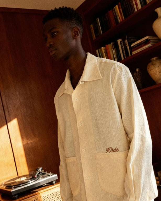 Hombre con sobrecamisa de algodón seersucker color crudo de KÖDE, de pie en un salón con luz cálida, frente a una estantería de madera.