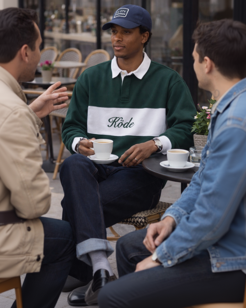 Modelo masculino negro sentado en una cafetería con polo rugby verde y blanco de KÖDE, escena cotidiana y estilo casual premium.